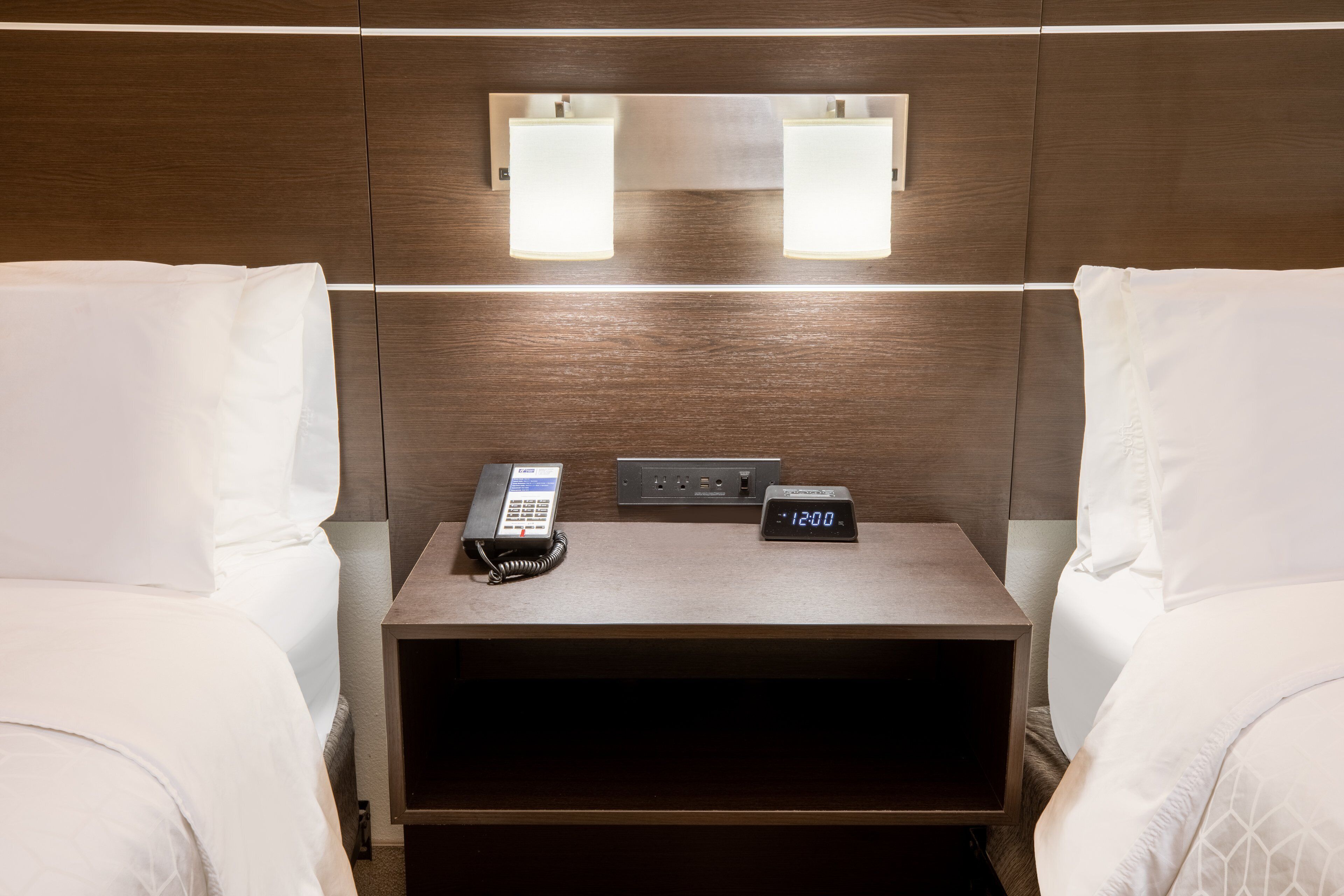 A hotel room with two beds, a bedside table with a phone and a digital clock, and wall-mounted lights.