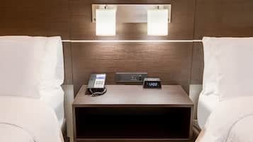 A hotel room with two beds, a bedside table with a phone and a digital clock, and wall-mounted lights.