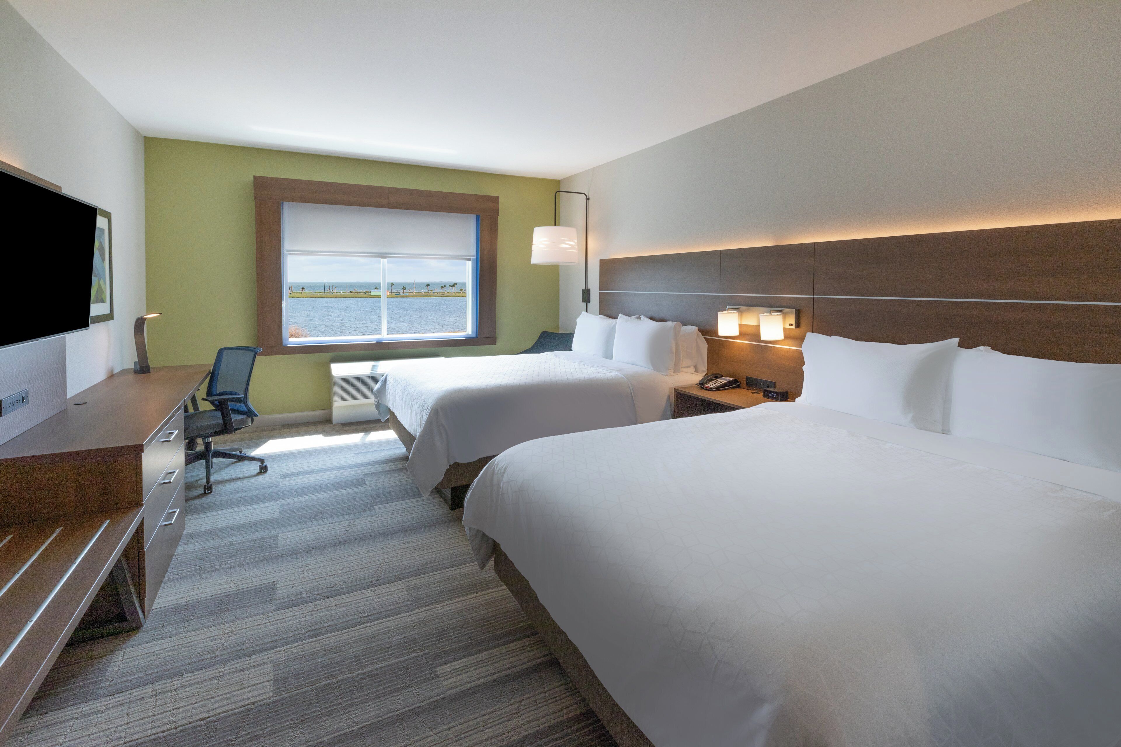 Habitación de hotel con dos camas, un escritorio con silla, televisor y ventana con vista al mar.