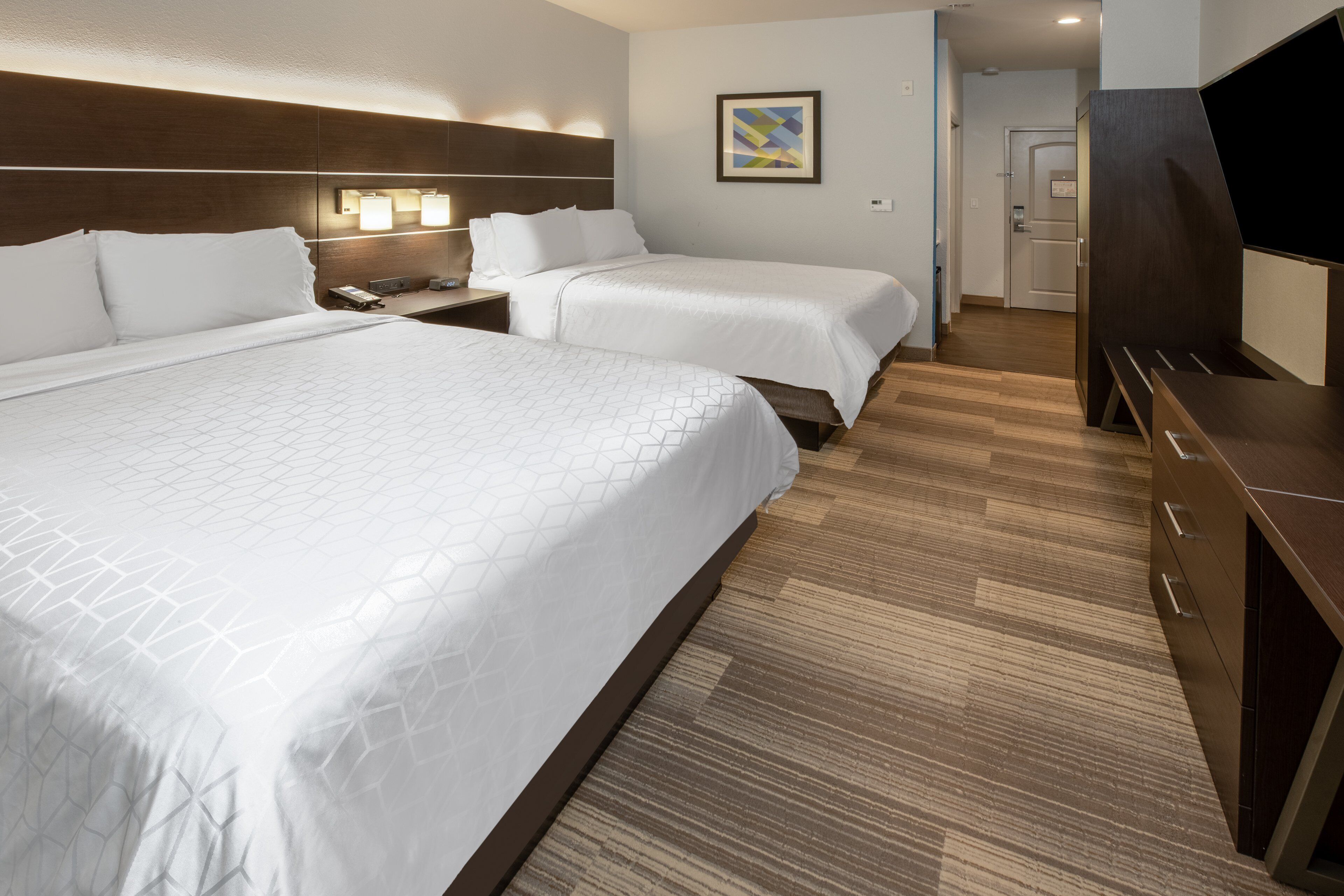 A hotel room with two beds, a wooden headboard, a bedside table with a lamp, and a TV.
