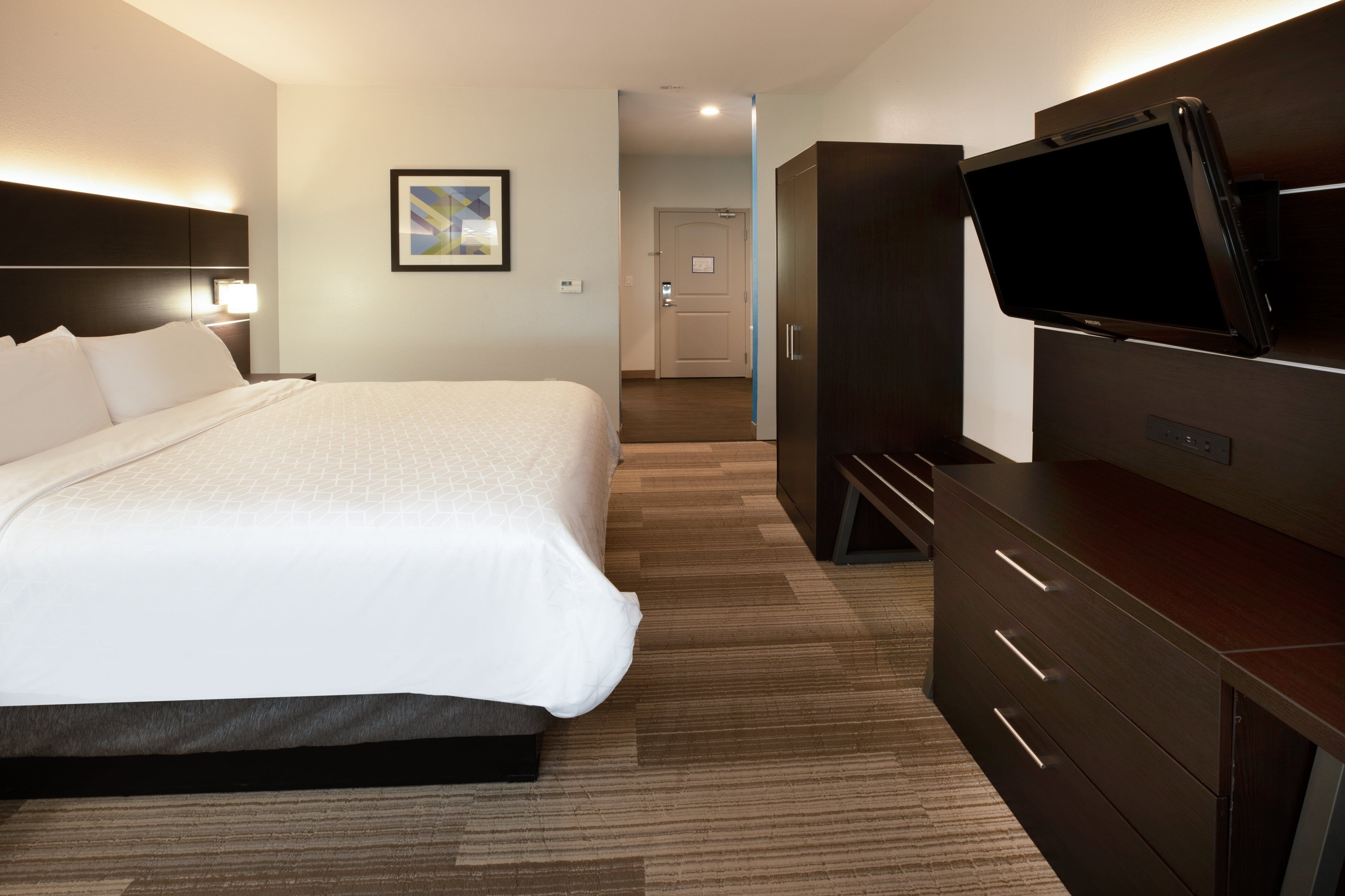 A hotel room with a large bed, a flat-screen TV mounted on the wall, a dresser, and a framed picture on the wall.