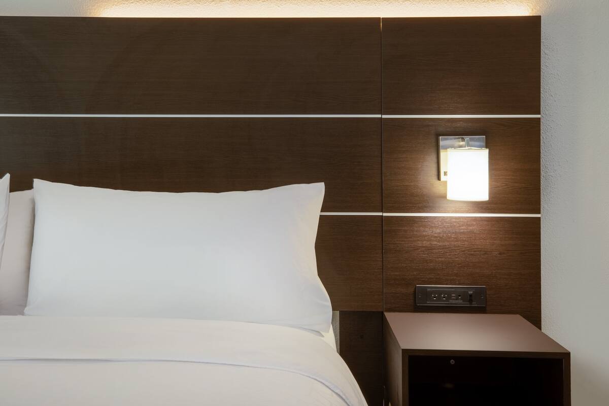 Una cama bien hecha con sábanas blancas, una mesita de noche fijada a la pared con una lámpara y un panel de control.