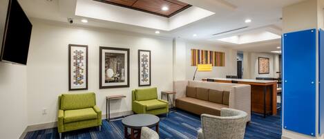Un moderno vestíbulo de hotel con alfombra azul, sillones verdes, un sofá color beige y una zona de comedor con bar.
