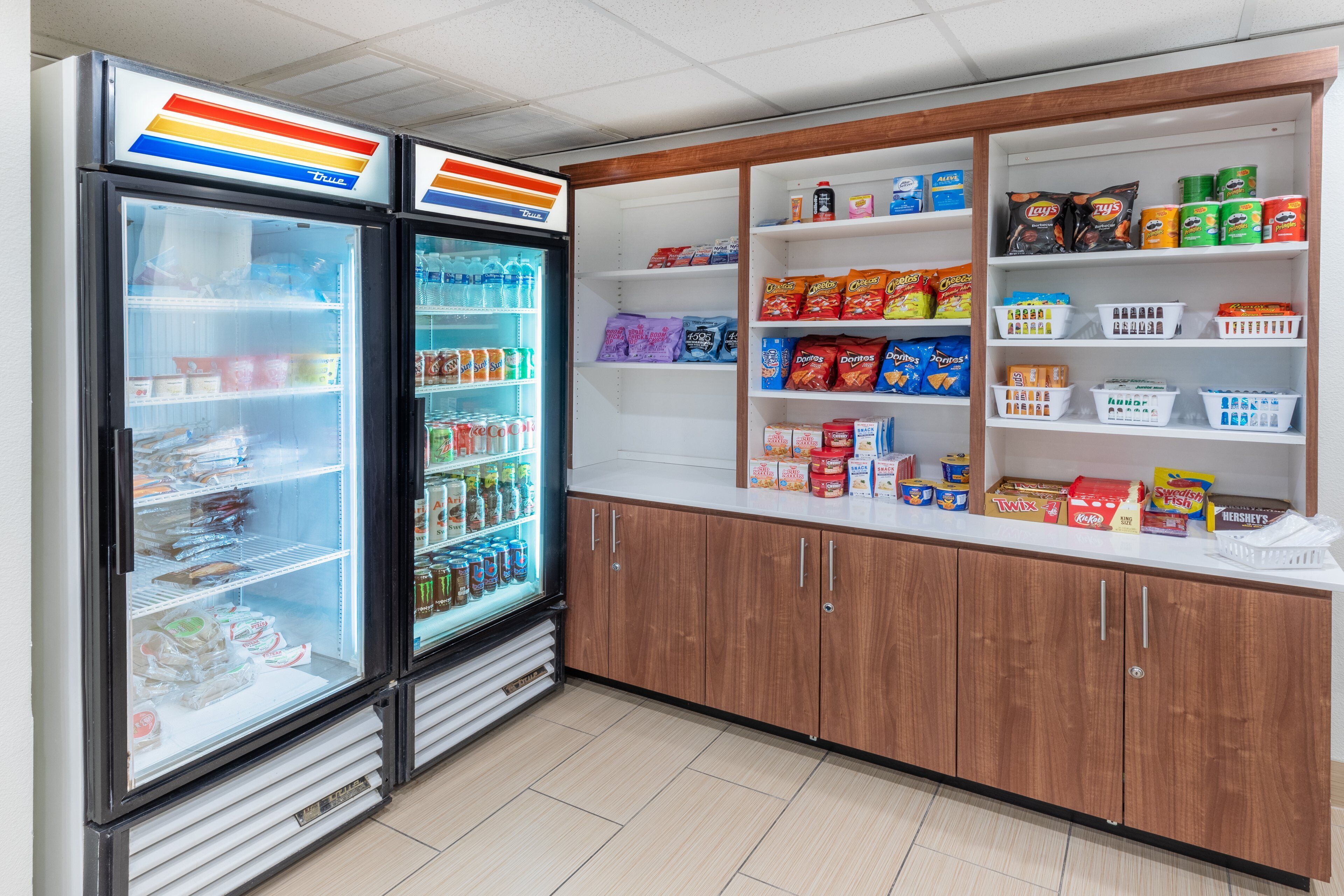 Un expositor refrigerado con diversas bebidas y aperitivos, armarios de madera con estantes llenos de bocadillos, y un piso de baldosas limpias.