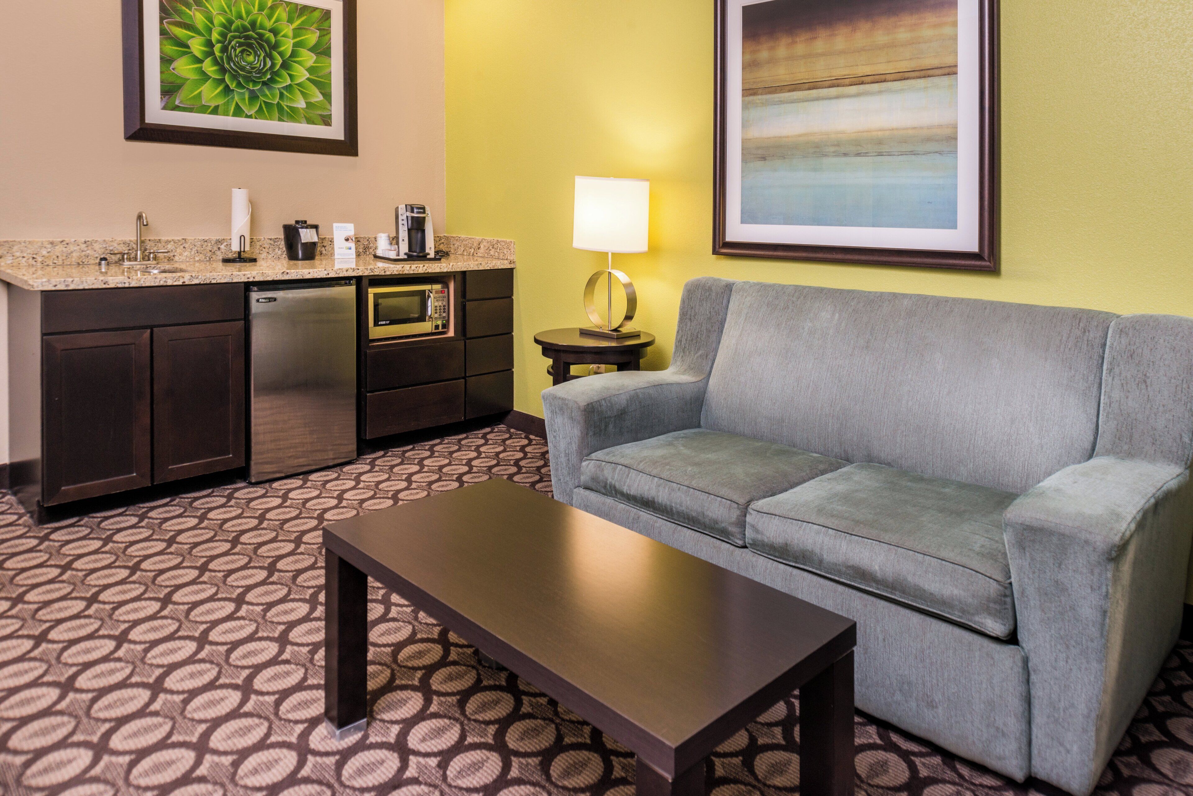 A hotel room with a kitchenette, a gray sofa, a coffee maker, a lamp, and a patterned carpet.