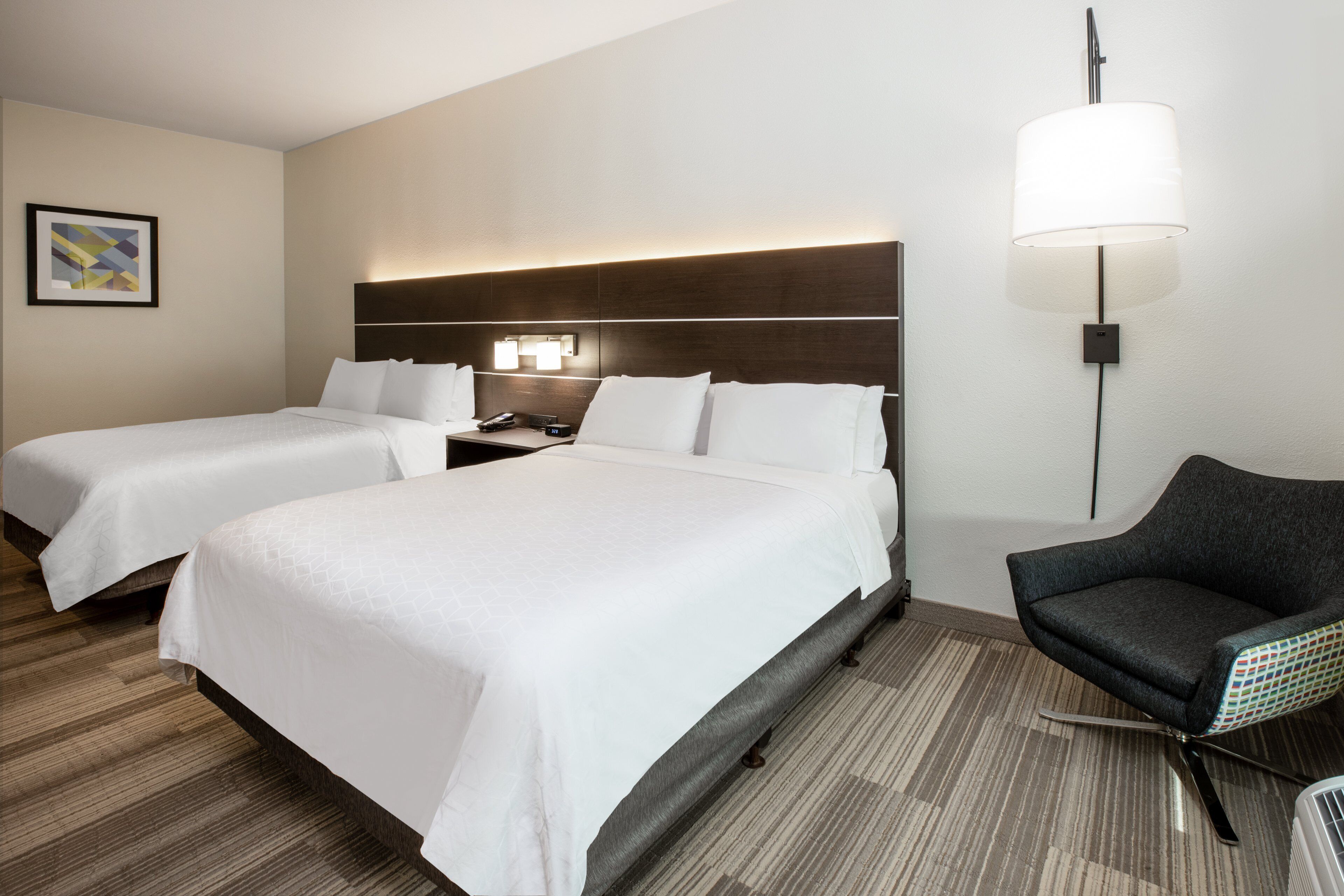 A hotel room with two beds, a nightstand, a lamp, and a chair.