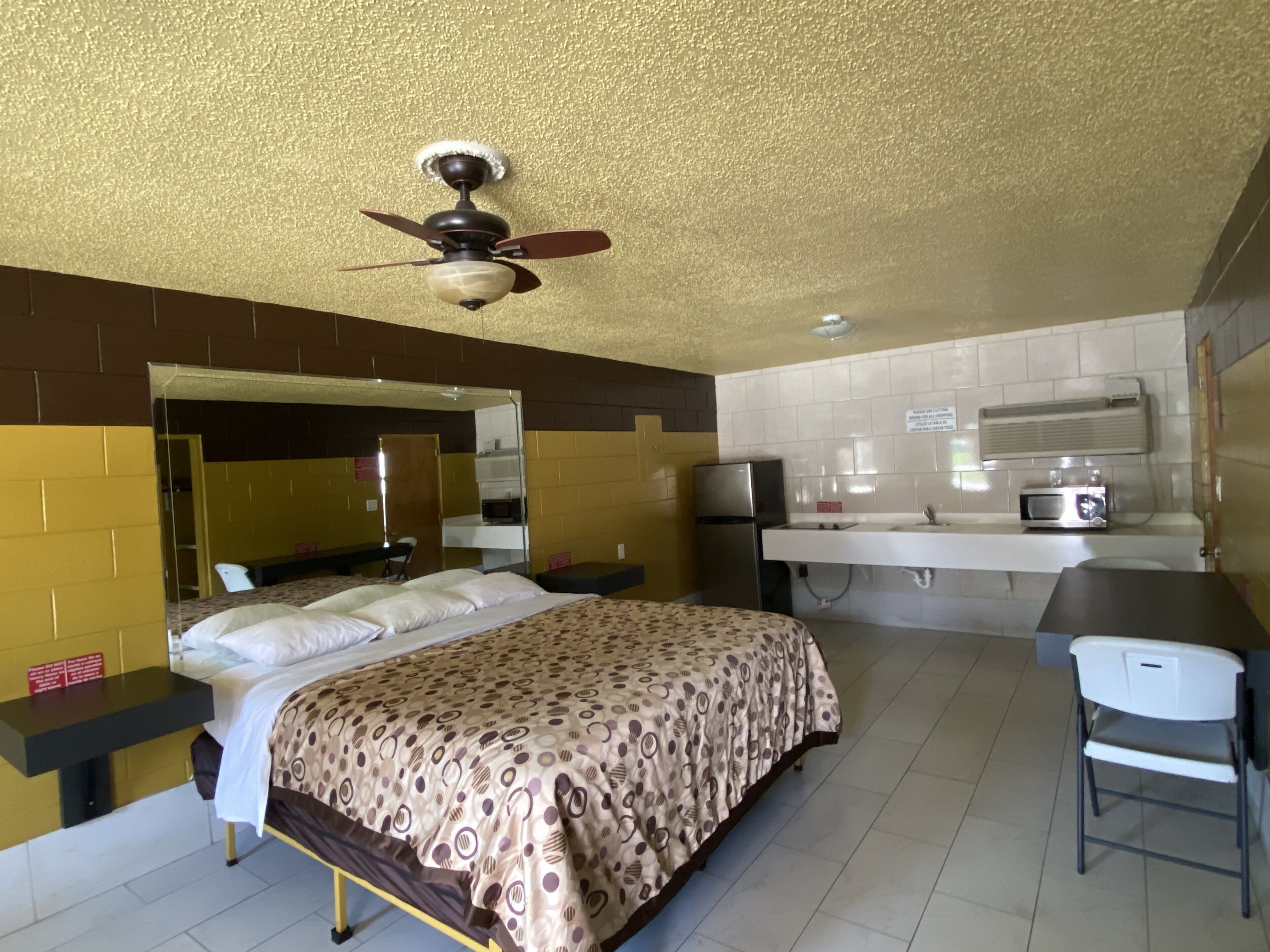 Quarto de hotel com cama, escrivaninha, cadeira, espelho, ventilador de teto, geladeira e micro-ondas.