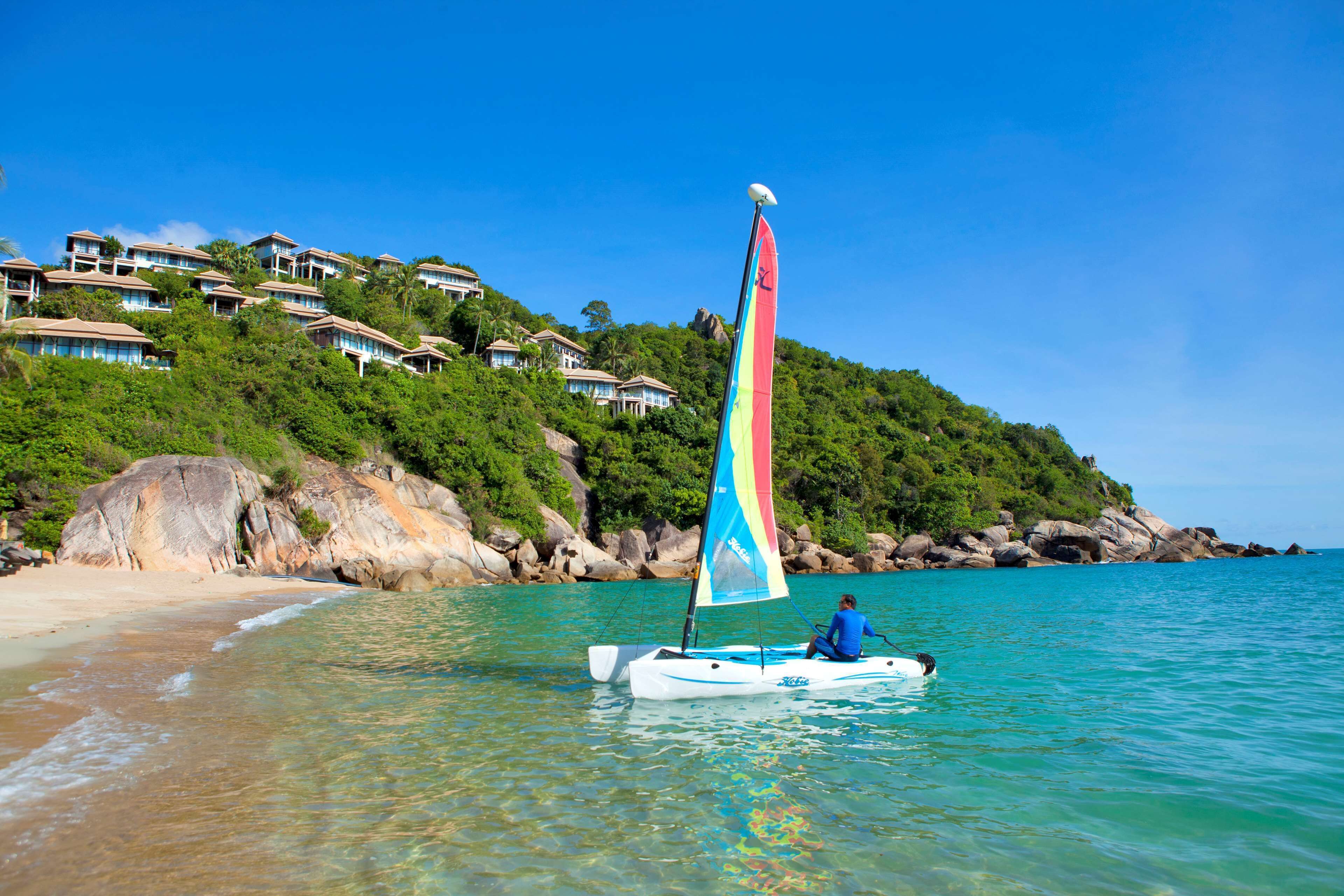A sailboat on clear turquoise water with a person on board, near a beach with houses on a hillside.
