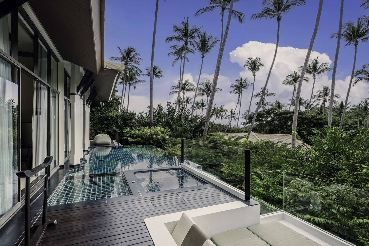 A modern outdoor infinity pool with a wooden deck and glass railings, surrounded by lush greenery and tall palm trees under a clear blue sky.