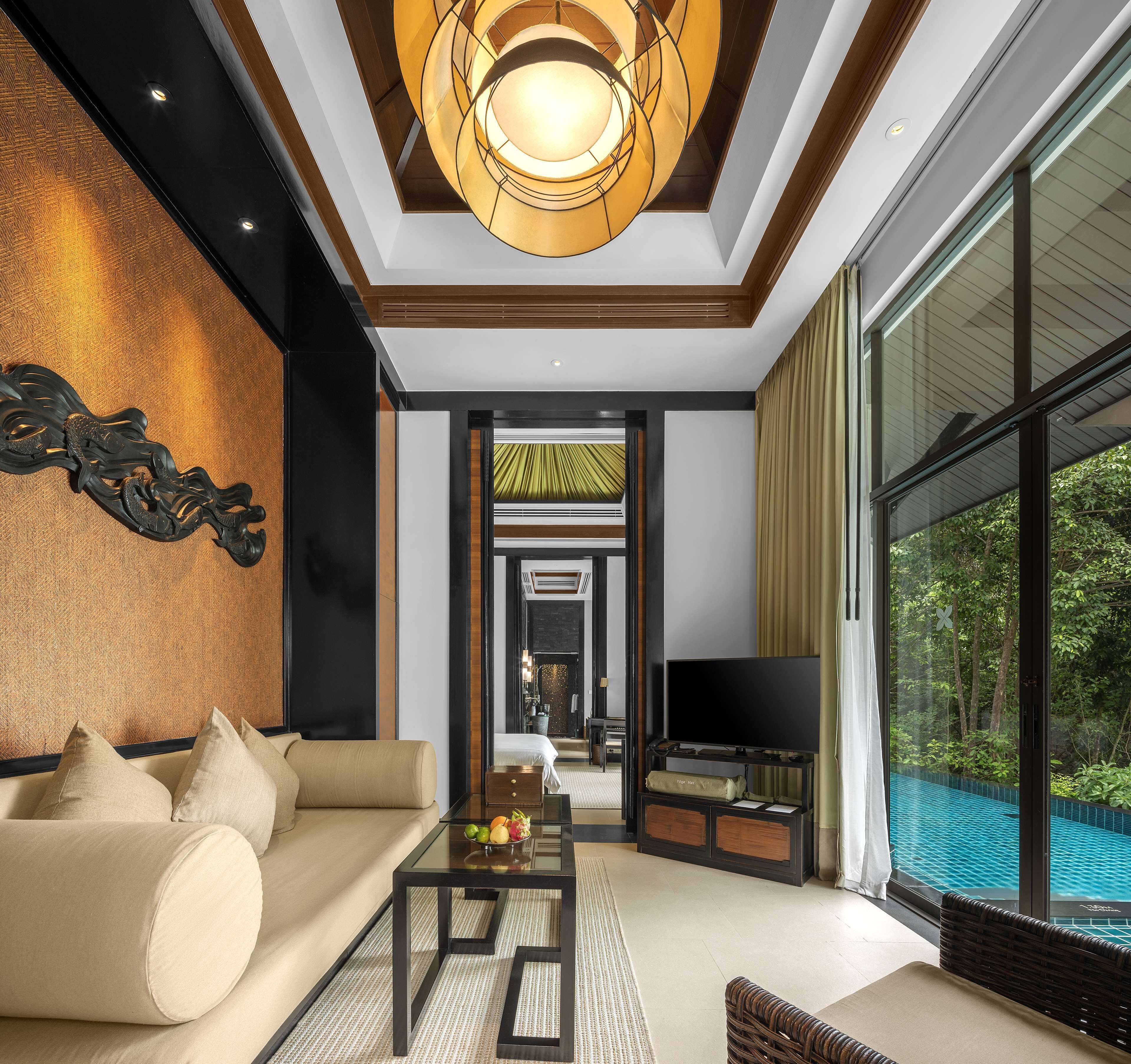 Un salon moderne avec un canapé, une table basse et un téléviseur, une grande fenêtre donnant sur une piscine et un plafond orné d’un luminaire original.