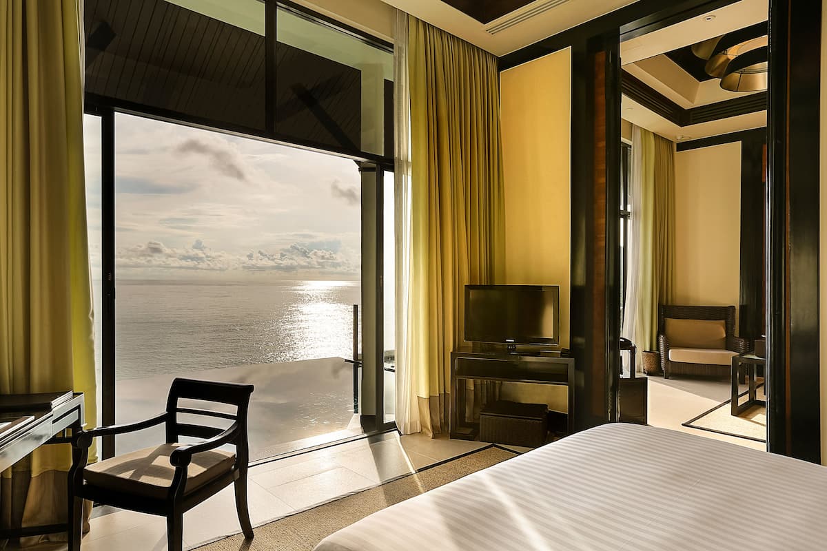 A hotel room with a large bed, a desk, a chair, a TV, and a balcony with a view of the water.