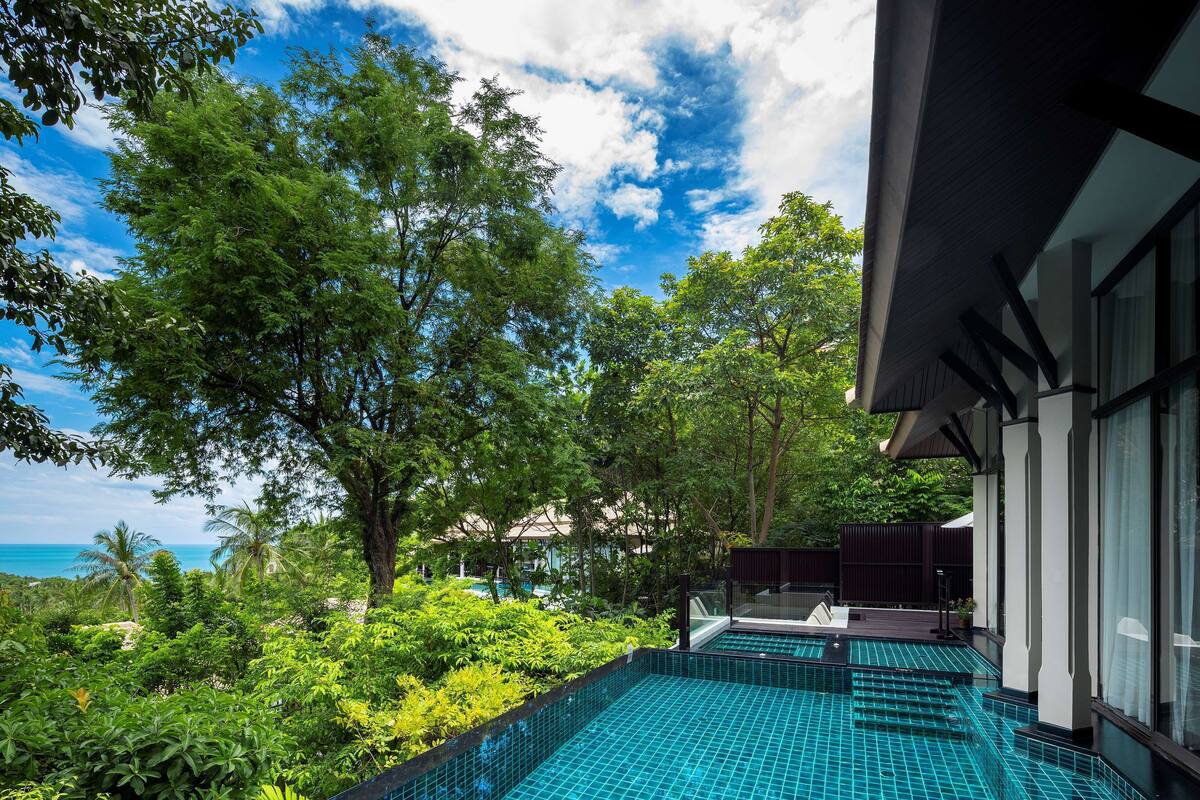 A swimming pool with a tiled floor, surrounded by lush greenery and trees, with a view of the ocean in the background.