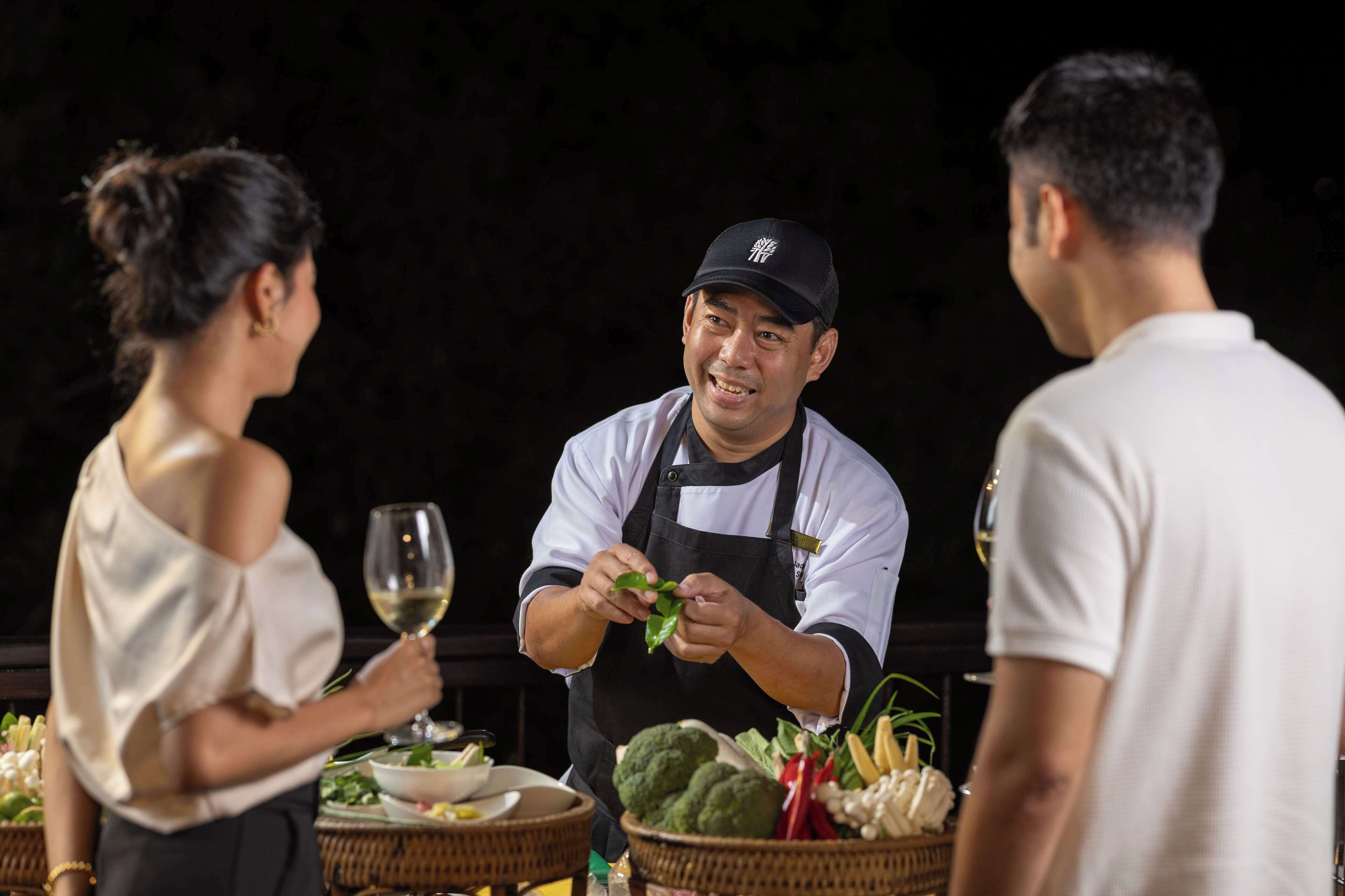 A person serving food to two guests at a table with a basket of vegetables.