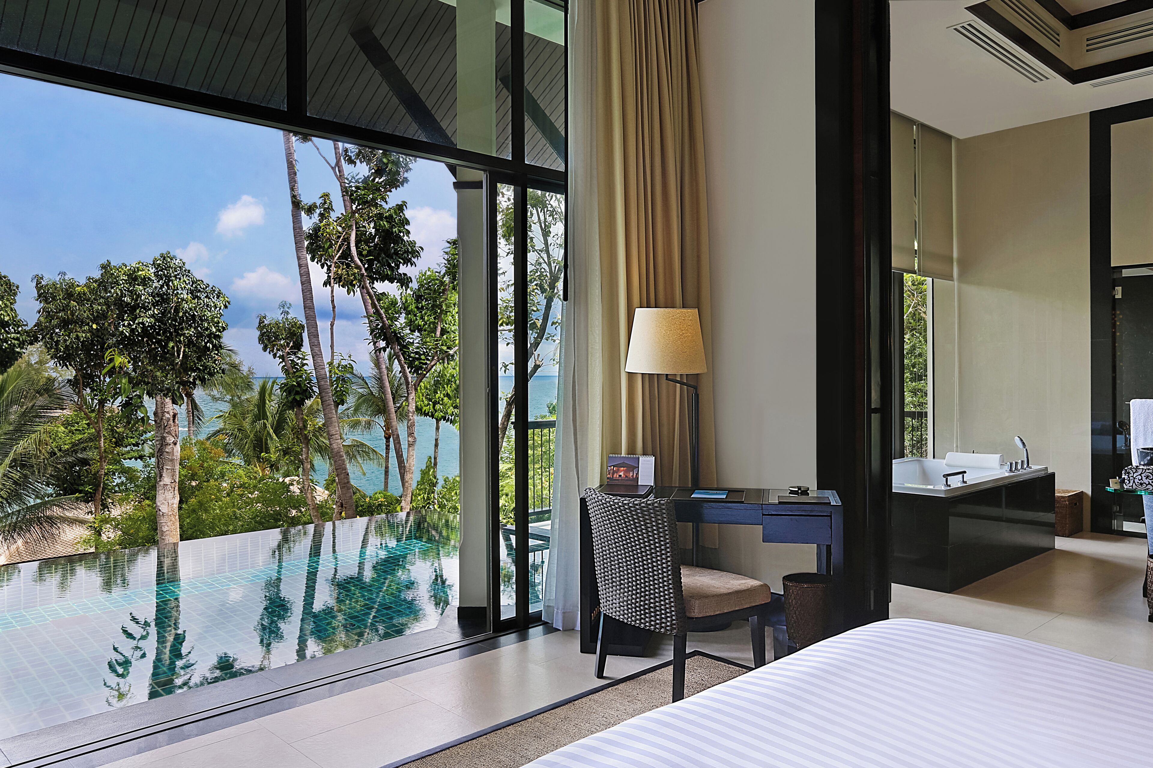 Une chambre d’hôtel dotée d’une grande fenêtre, d’un bureau, d’une chaise, d’un lit et d’une salle de bain avec un lavabo et un miroir.
