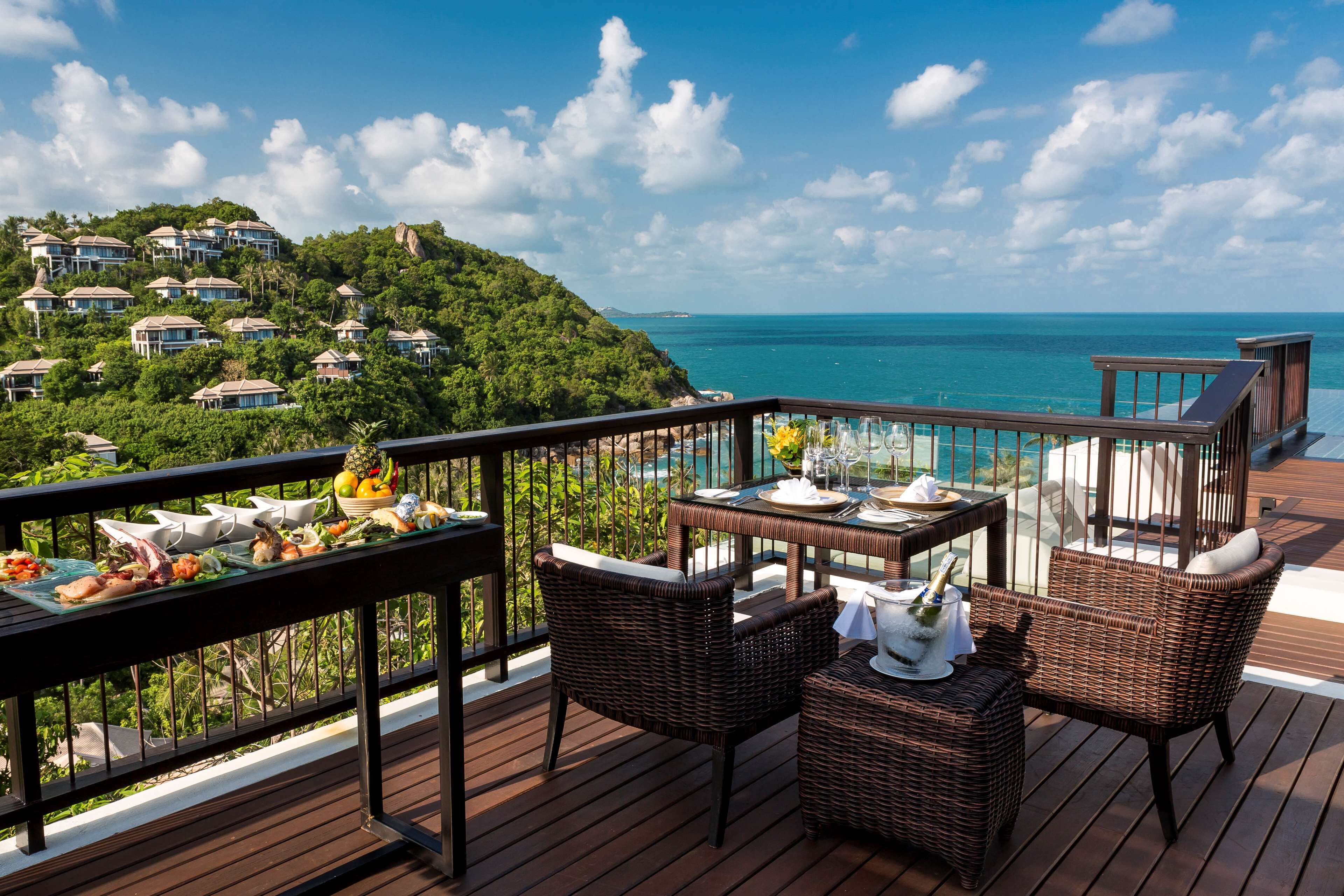 A deck with wicker furniture, a table set for dining, and a view of the ocean and hillside residences.