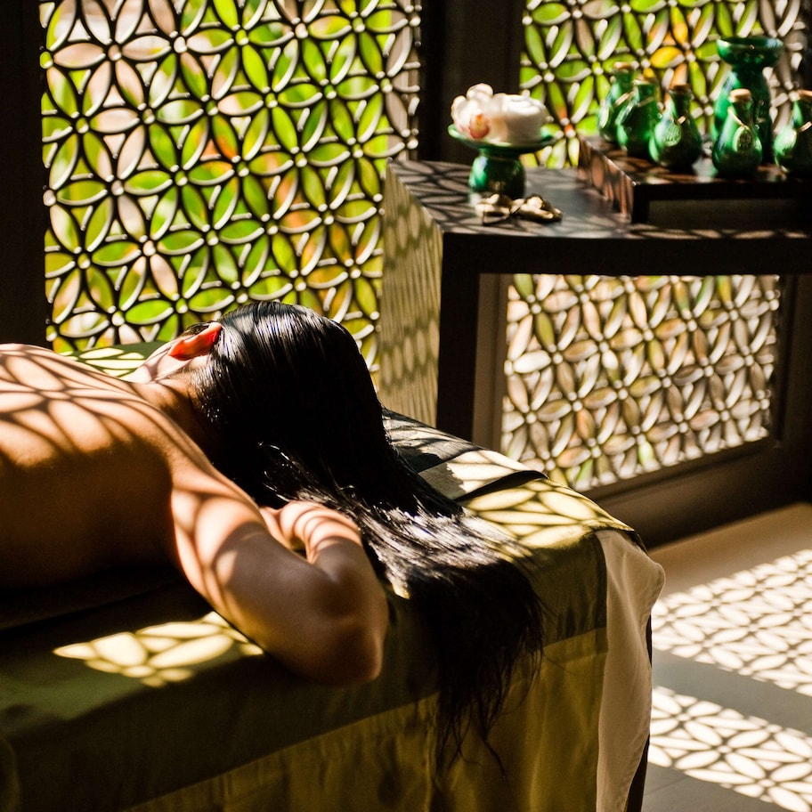 A person lying on a massage table in a room with a patterned window and a small table with decorative items.