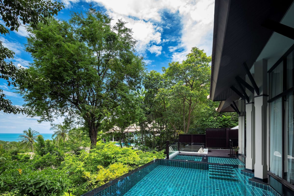 A swimming pool with a tiled floor, surrounded by lush greenery and trees, with a view of the ocean in the background.