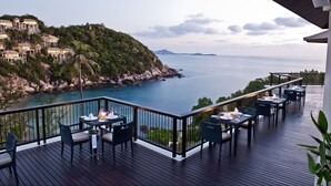 A deck with tables and chairs overlooking a coastal area with a rocky shoreline and a view of a distant mountain range. - Banyan Tree Samui (Koh Samui)