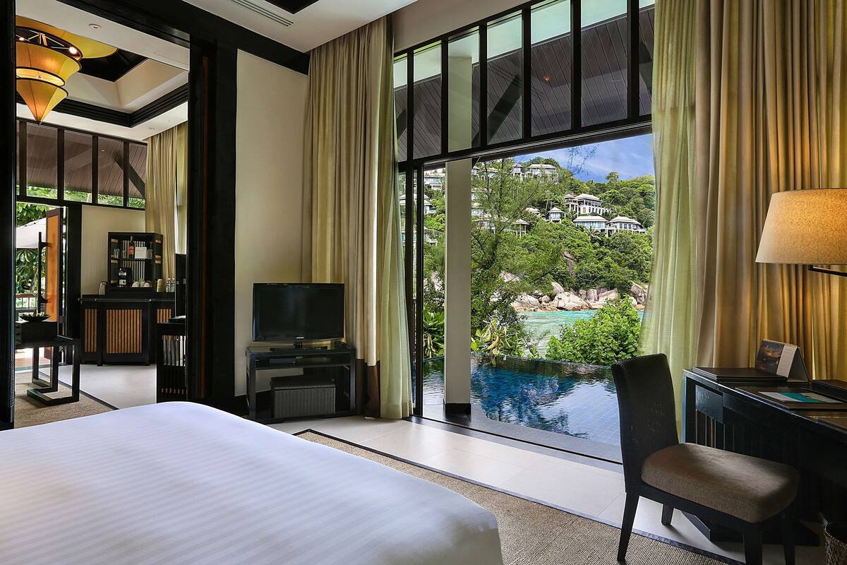 A hotel room with a large bed, a desk with a lamp, a TV, and a view of a swimming pool and greenery.