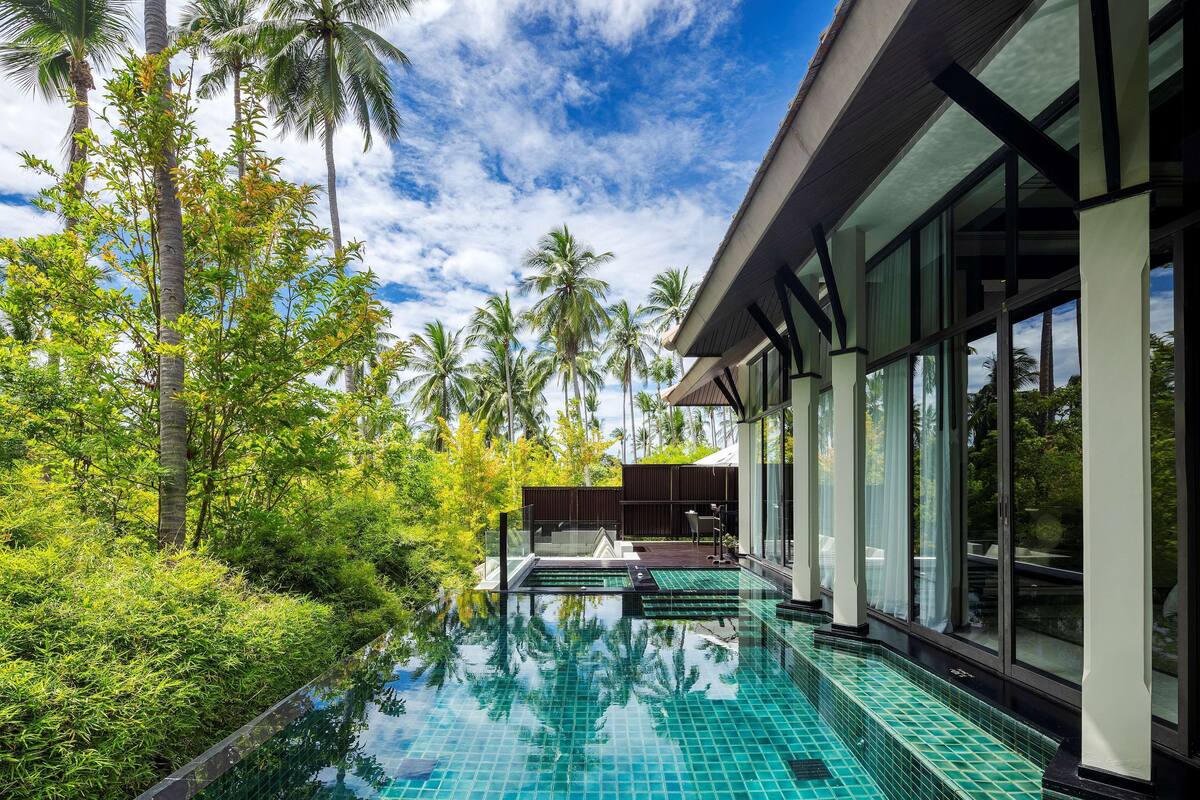 A modern pool area with clear blue tiles, surrounded by lush greenery and tall palm trees.