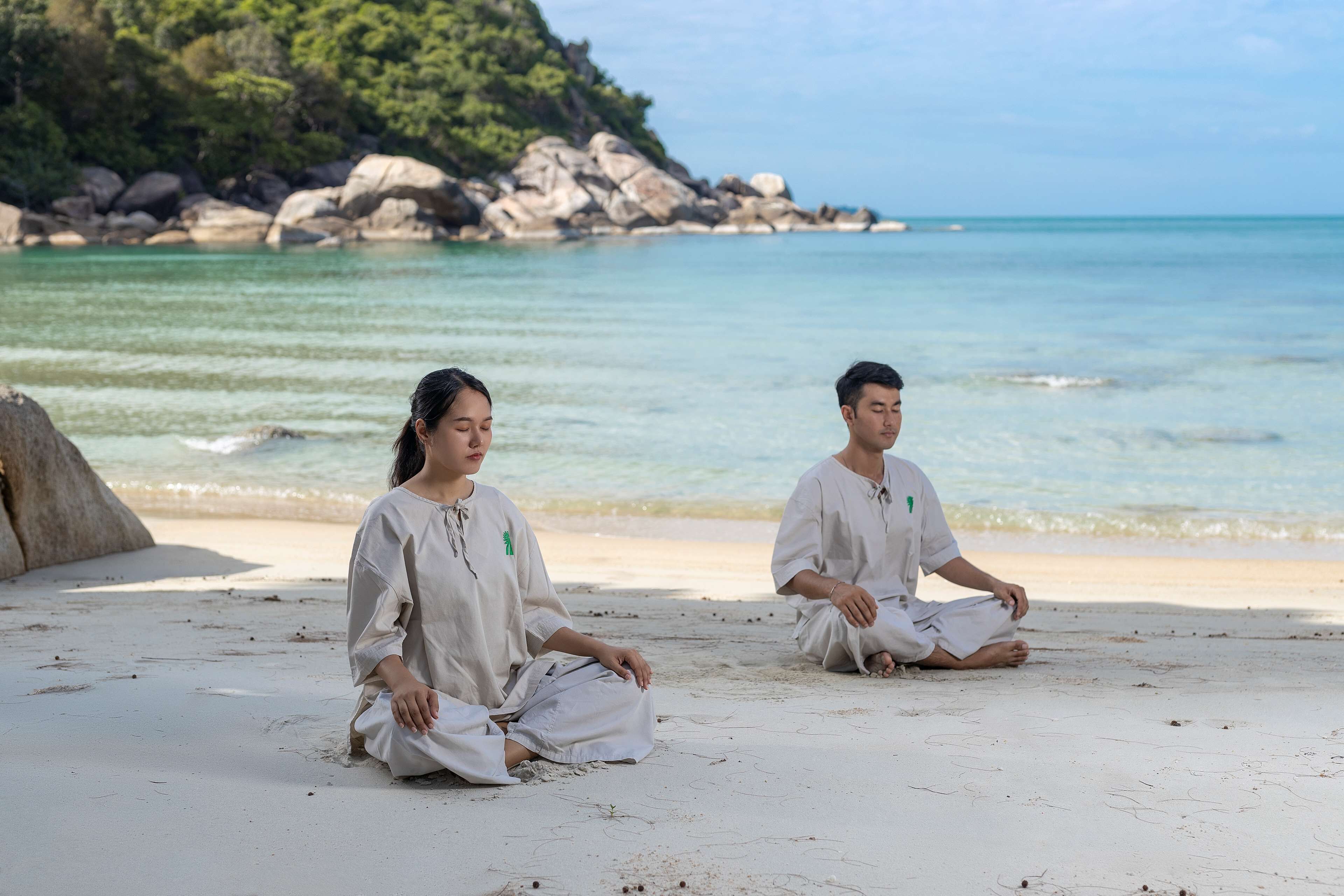 Two individuals sitting on a sandy beach with a rocky shoreline and calm sea in the background.
