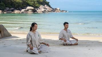 Two individuals sitting on a sandy beach with a rocky shoreline and calm sea in the background.