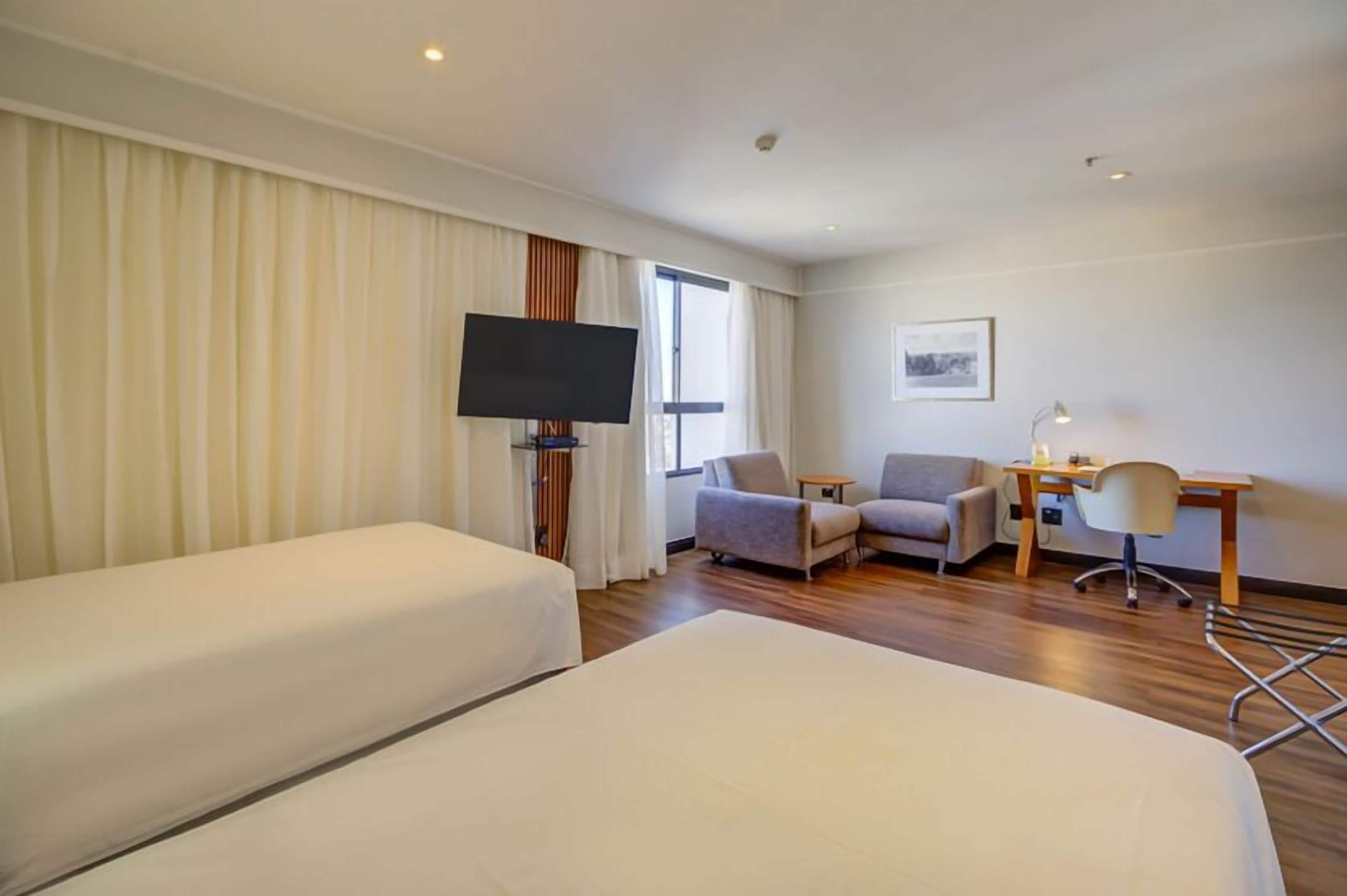 A hotel room with two beds, a desk, a chair, a TV, and a window with curtains.