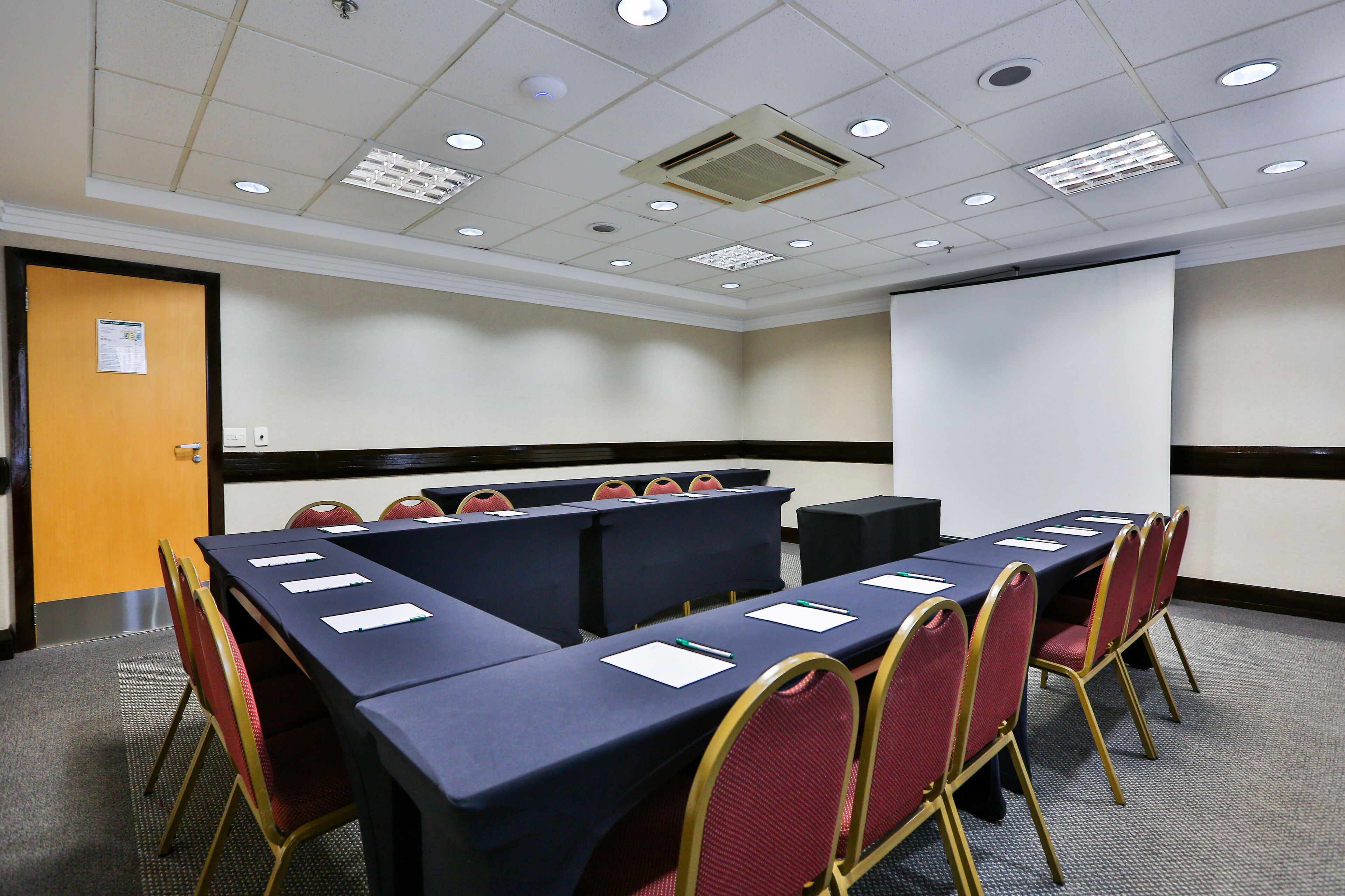 A conference room with a long table, red chairs, a projector screen, and a door with a sign.