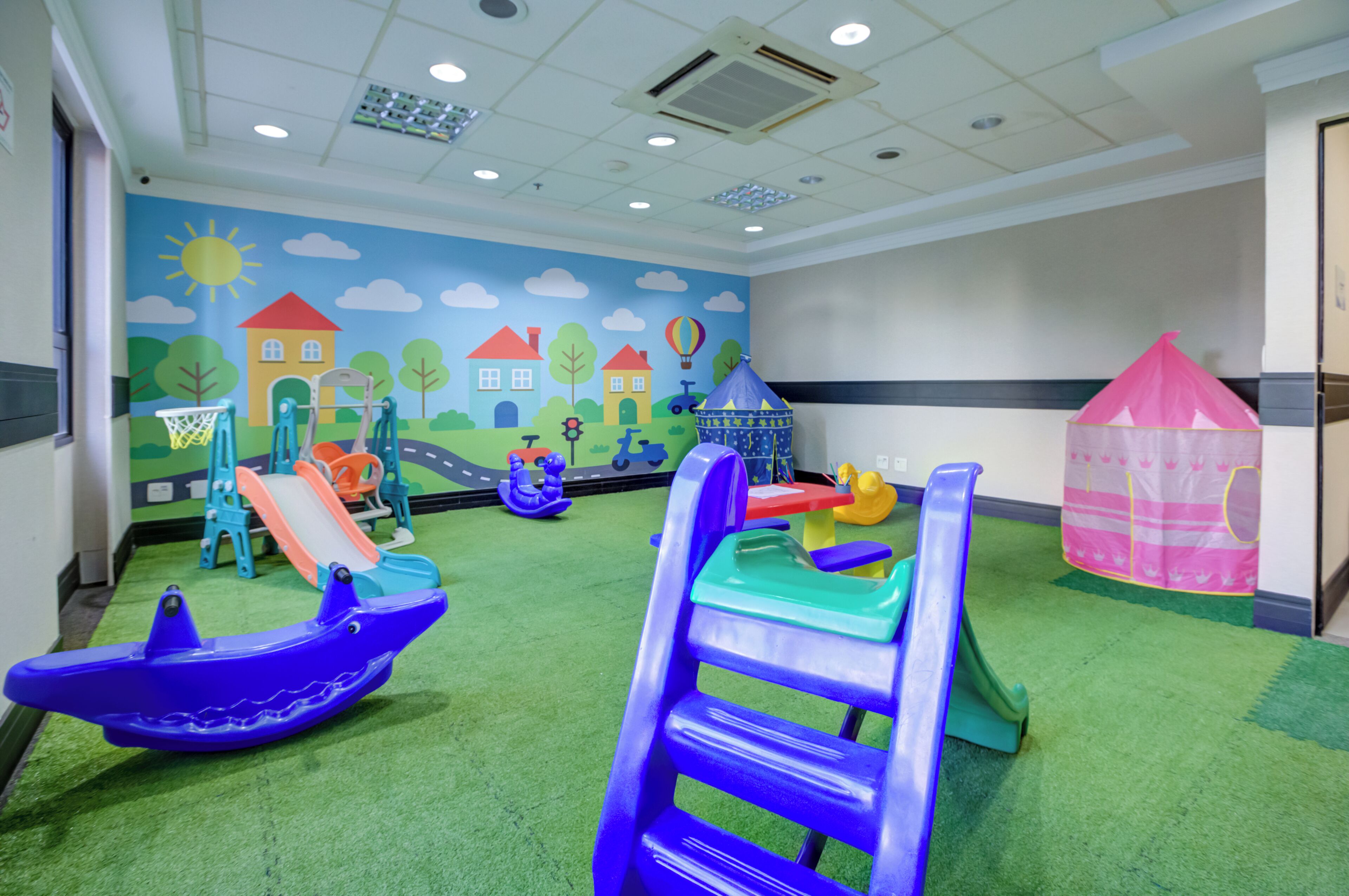 A children's play area with various play structures, including slides and climbing frames, on a green carpeted floor.