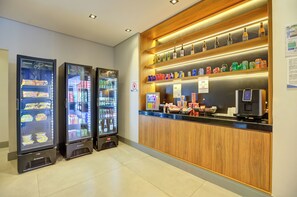 Zona de recepción de un hotel con mostrador de madera, dispensador de bebidas y máquina expendedora de bebidas.