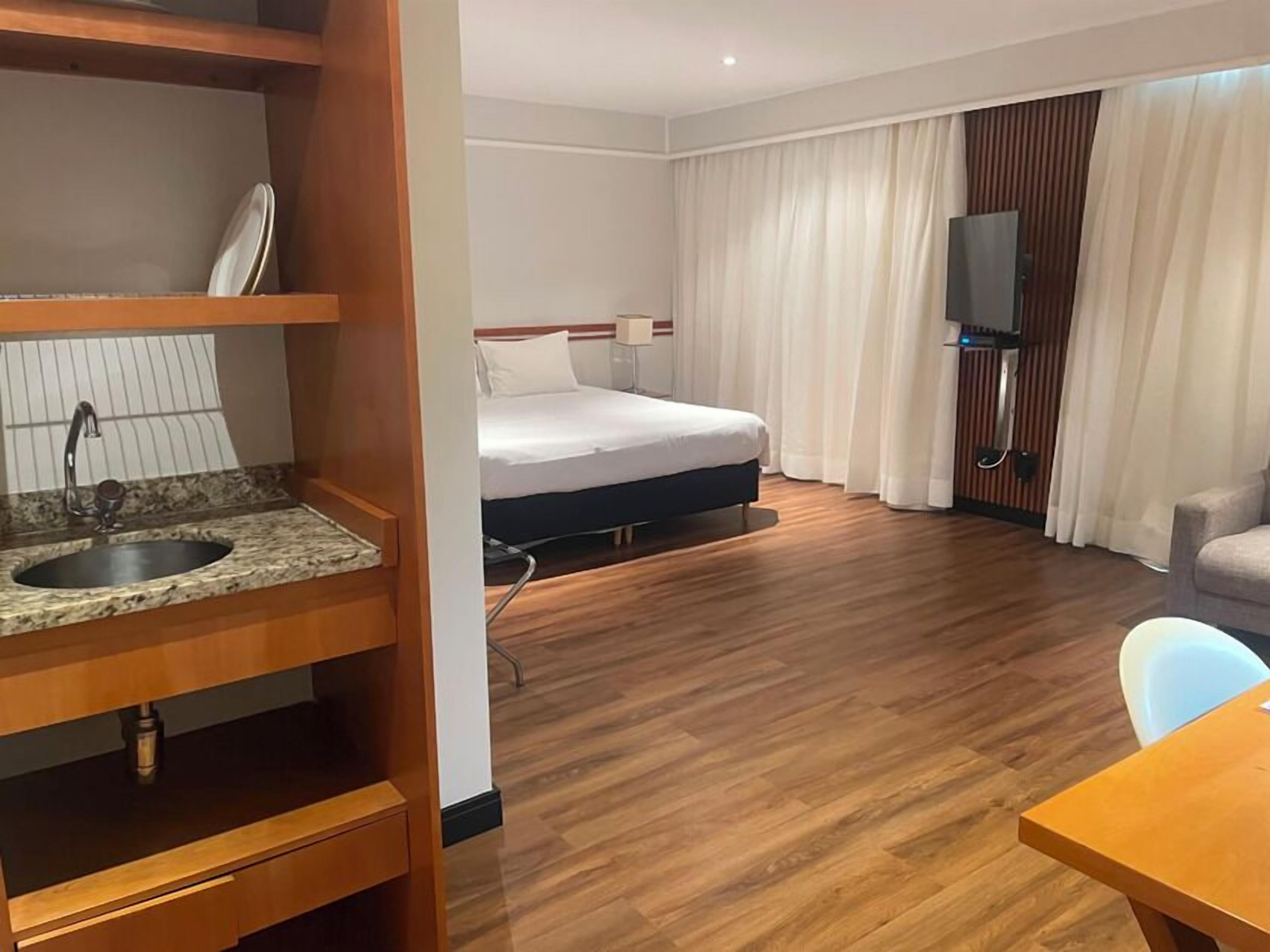 A hotel room with a bed, a television, a wooden floor, a sink, and a chair.