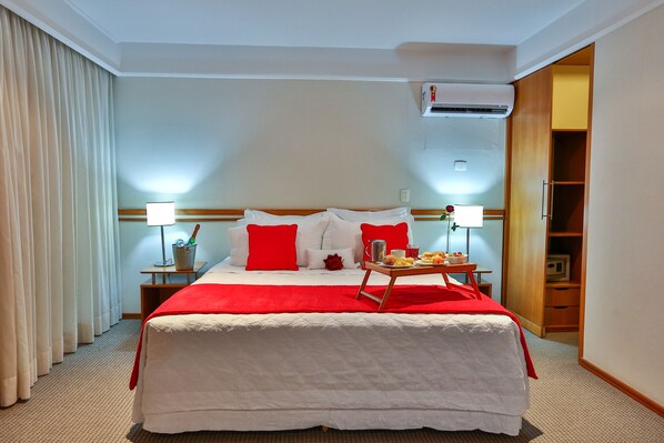Habitación de hotel con una cama grande, ropa de cama roja y blanca, dos lámparas de noche, una mesita de noche con lámpara, una pequeña mesa con artículos de desayuno y un armario.