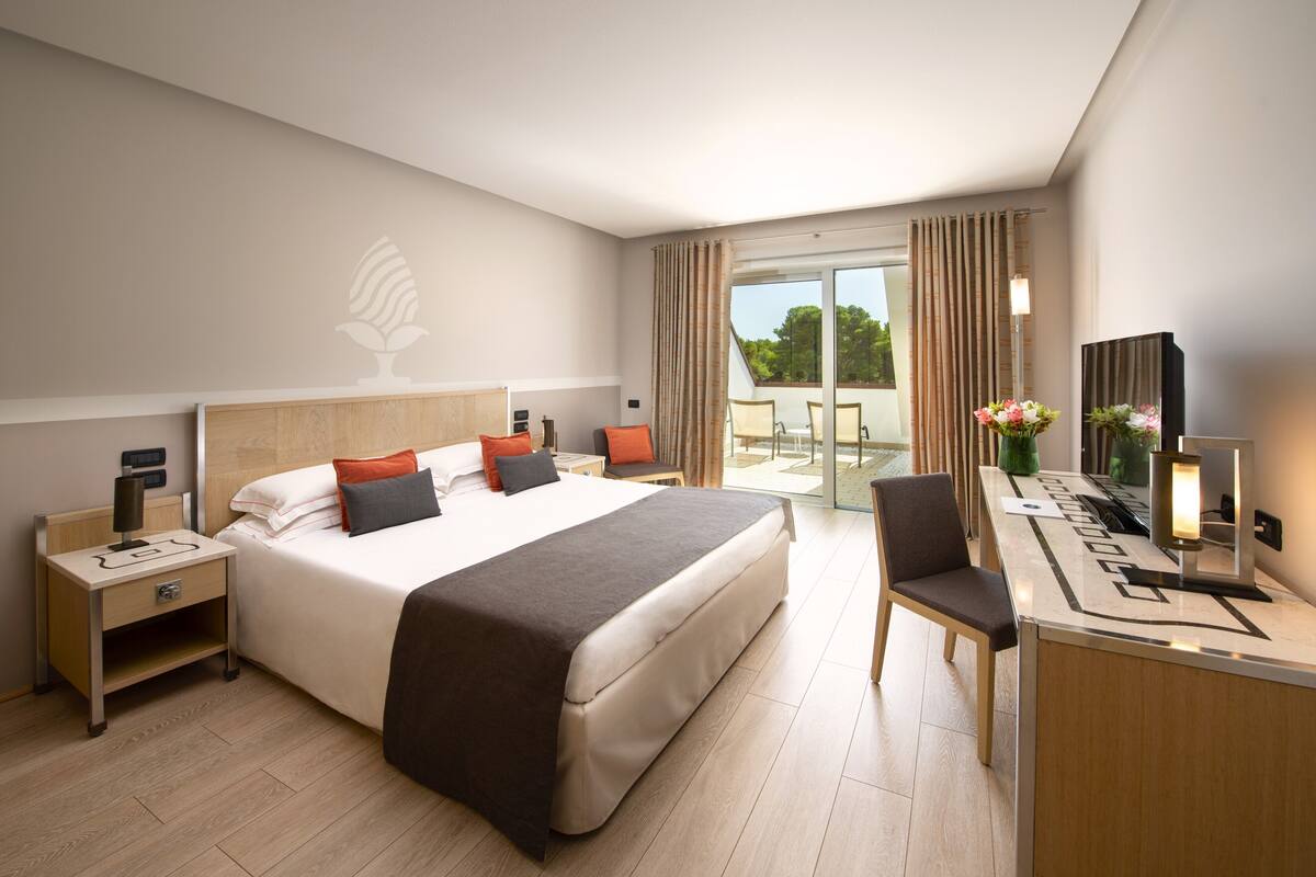 Habitación de hotel con cama, escritorio, silla, mesita de noche, lámpara, espejo y vistas al exterior.