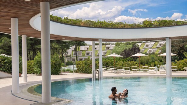 Una piscina con borde curvo, rodeada de columnas blancas y un área techada con techo de madera.