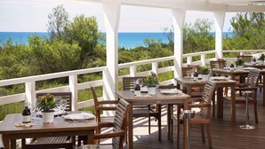Un área de comedor al aire libre con mesas y sillas de madera, barandas blancas y vista al océano.