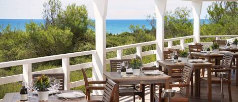 An outdoor dining area with wooden tables and chairs, white railings, and a view of the ocean.