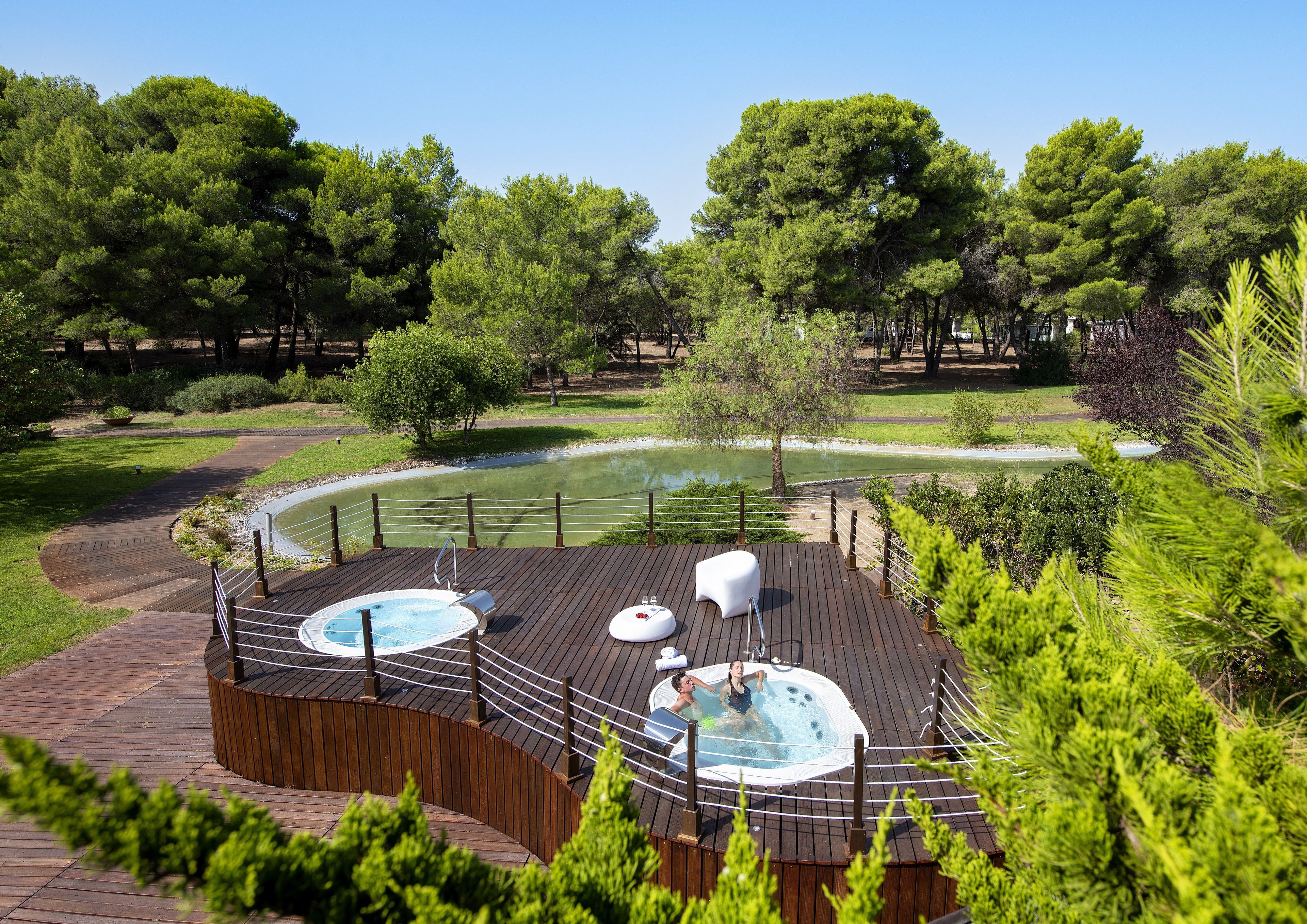Un área de hidromasaje con dos jacuzzis y un espacio de descanso en una terraza de madera, rodeado de vegetación exuberante y un estanque.