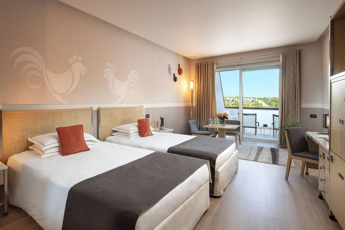 Habitación de hotel con dos camas, un escritorio, una silla y vistas al exterior.