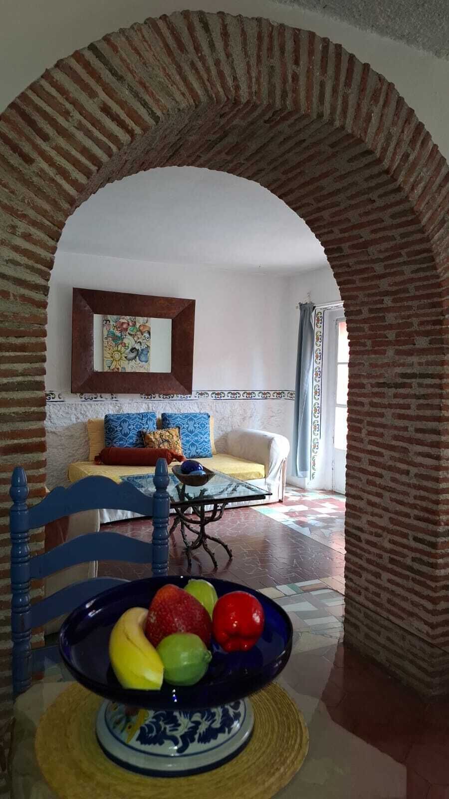 Un comedor con una mesa, sillas y un frutero. Un arco conduce a una sala de estar con un sofá y un cuadro en la pared.