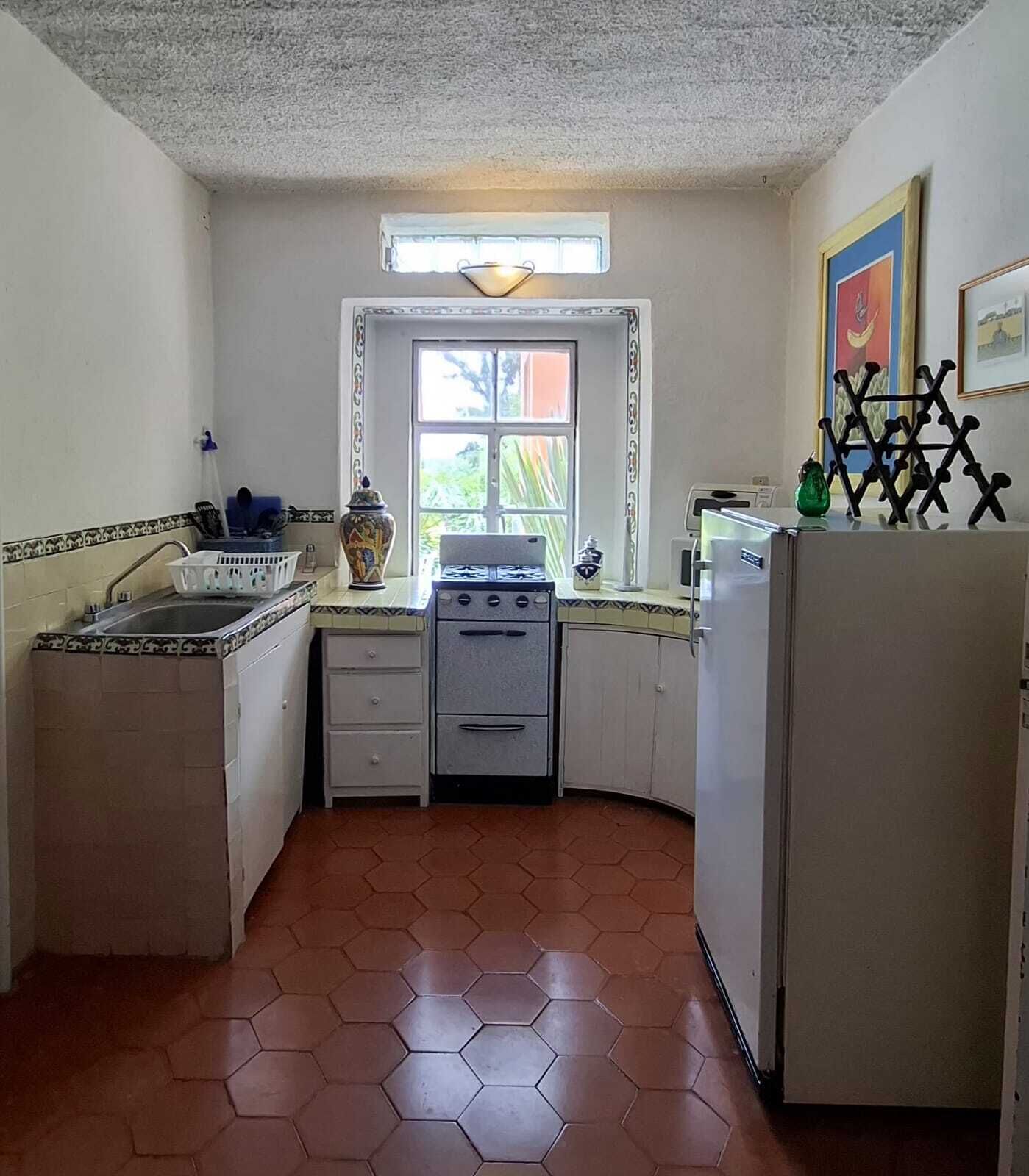 Una cocina con estufa, lavamanos y refrigerador, una ventana con cortinas y piso de baldosas.