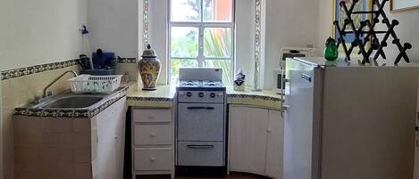 Una cocina con estufa, lavamanos y refrigerador, una ventana con cortinas y piso de baldosas.