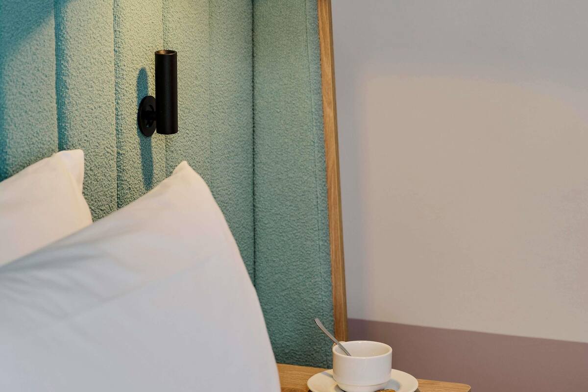 A hotel room with a bed, pillows, a bedside table with a cup and cookies, and a wall-mounted phone.