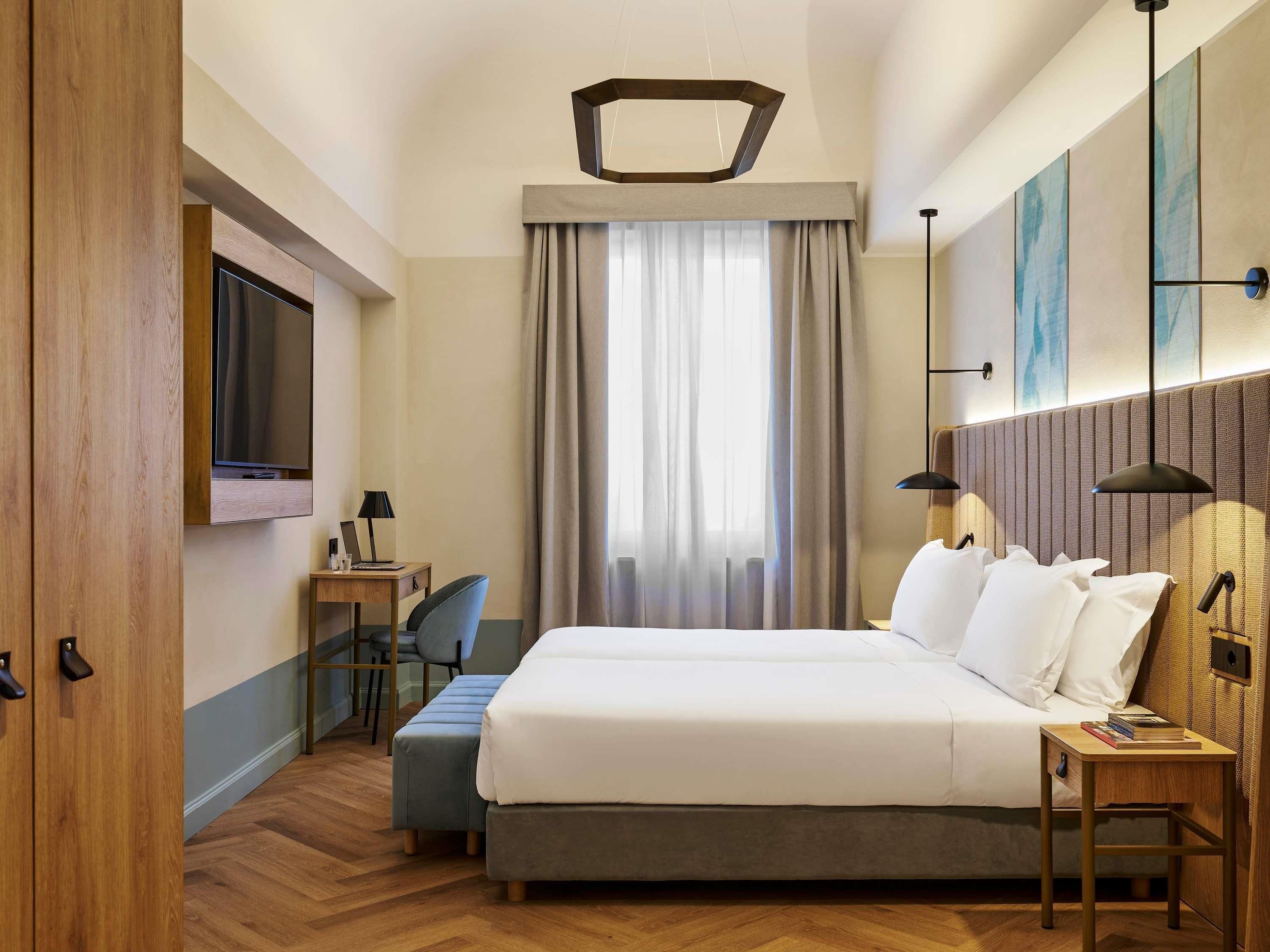Una camera d'albergo con un letto, una scrivania, una sedia, una TV e una finestra con tende.