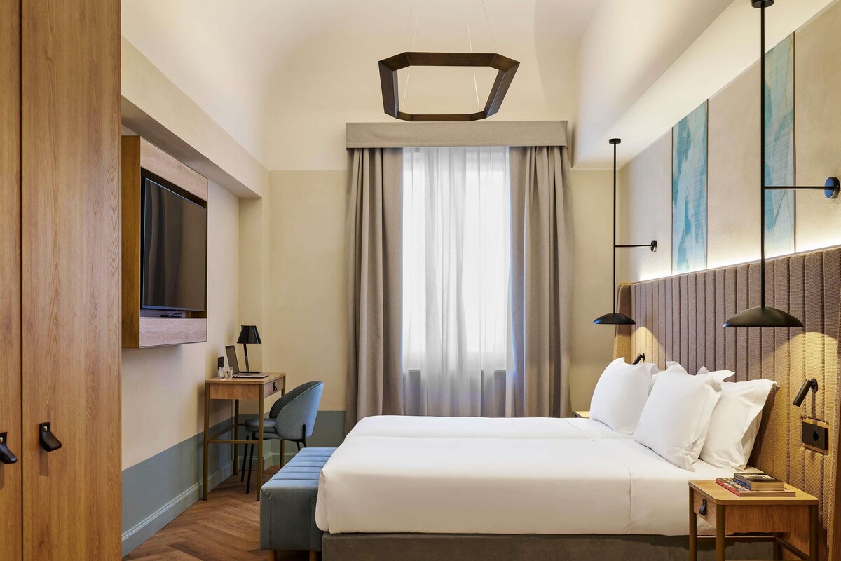 Une chambre d’hôtel avec un lit, un bureau, une chaise, une télévision et une fenêtre avec des rideaux.