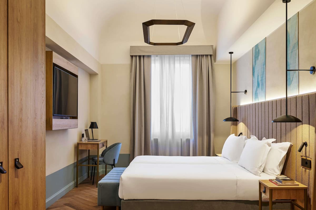 A hotel room with a bed, a desk, a chair, a TV, and a window with curtains.