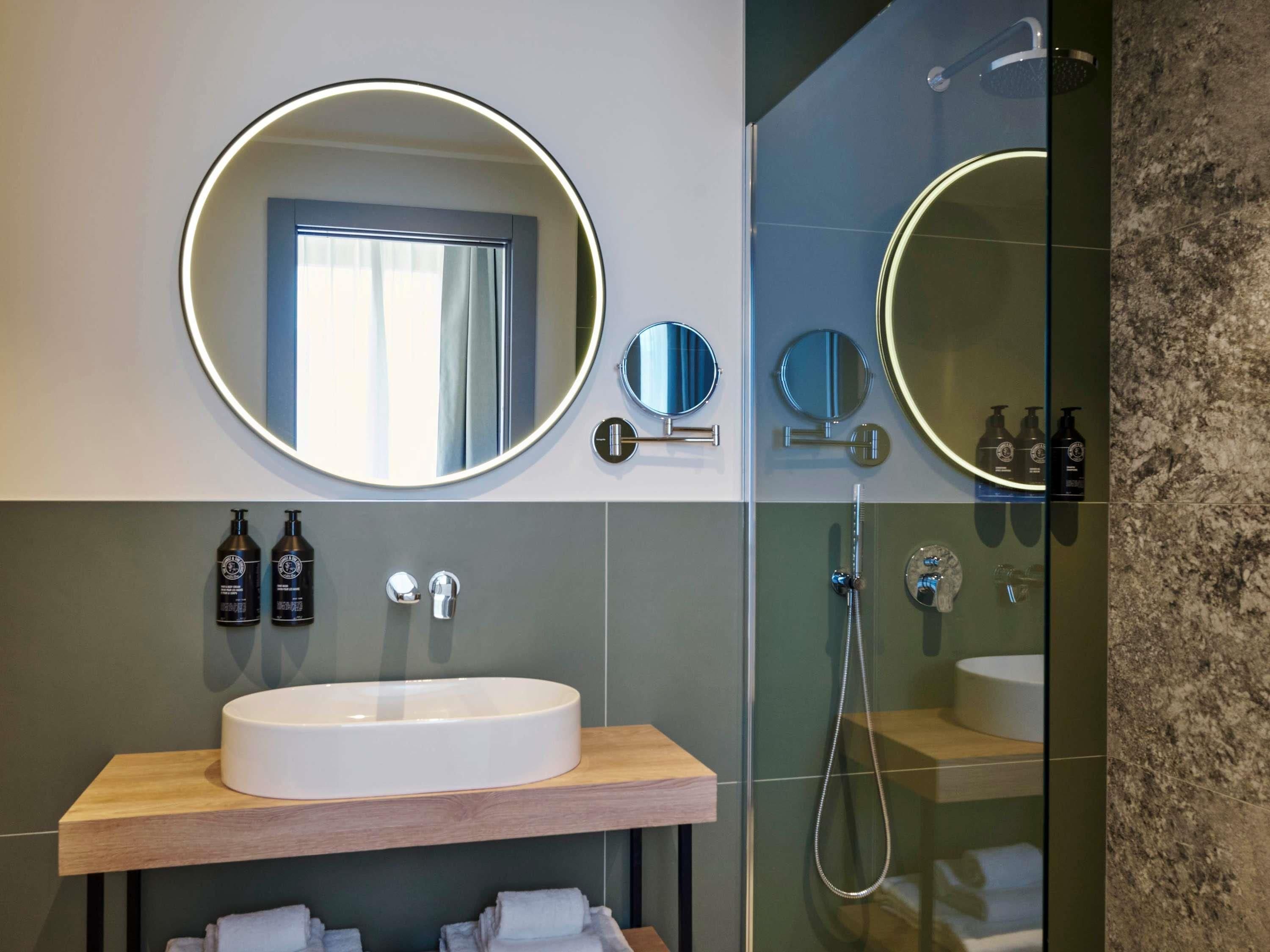 A modern bathroom with a large round mirror, a sink, and a glass-enclosed shower.
