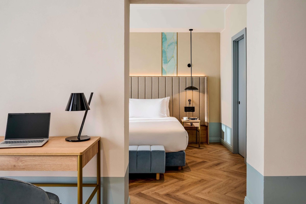 A hotel room with a bed, a desk with a laptop, a lamp, a bedside table with a cup and books, and a large window.