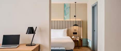 A hotel room with a bed, a desk with a laptop, a lamp, a bedside table with a cup and books, and a large window.
