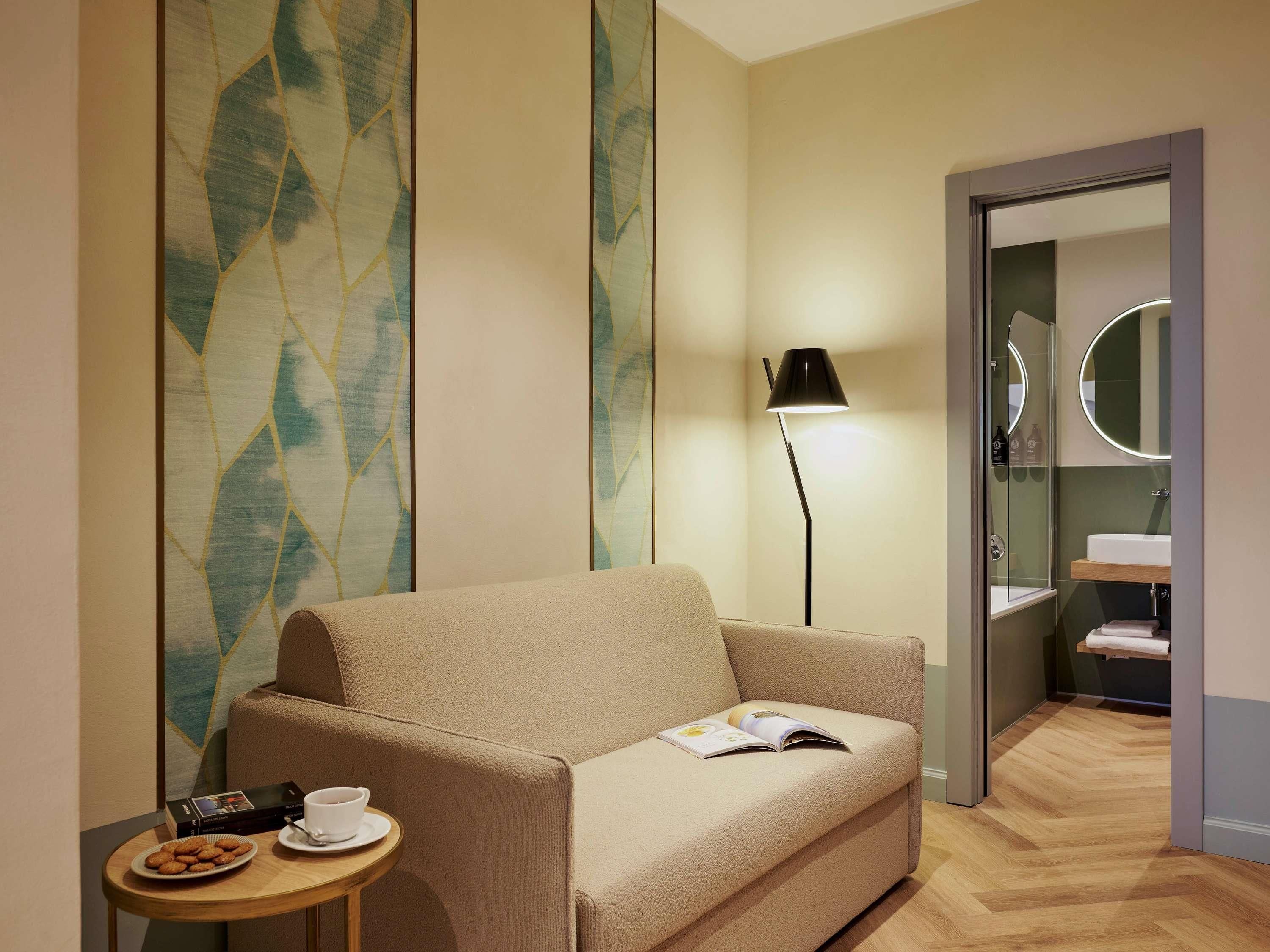 Una habitación de hotel moderna con un sofá color crema, una mesita redonda pequeña con una taza y galletas, una lámpara de pie y una zona de ducha con mampara de cristal.