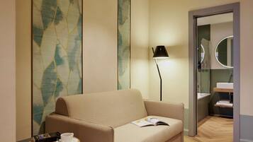 Una habitación de hotel moderna con un sofá color crema, una mesita redonda pequeña con una taza y galletas, una lámpara de pie y una zona de ducha con mampara de cristal.